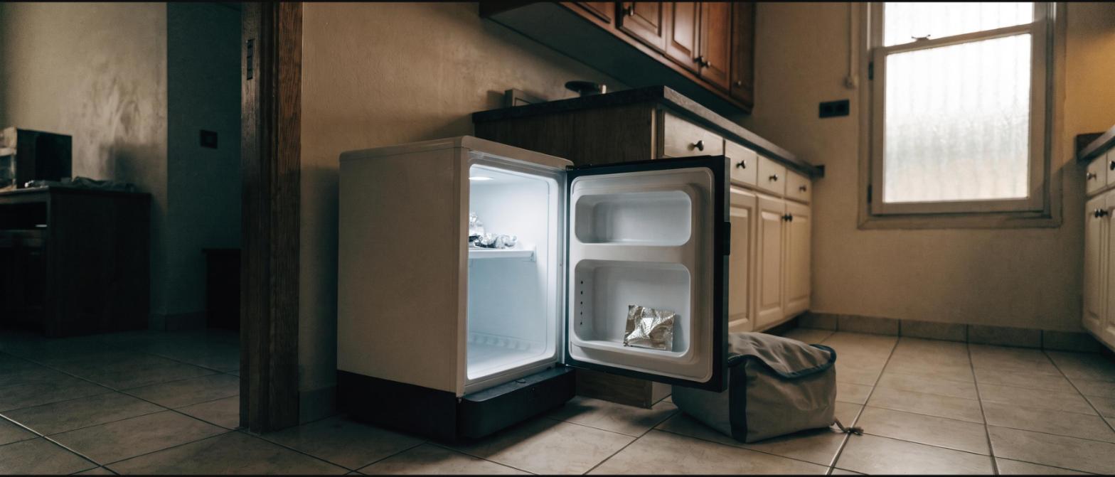 A small under-counter fridge standing open and nearly empty in a rental apartment kitchen with a soft cooler bag beside it