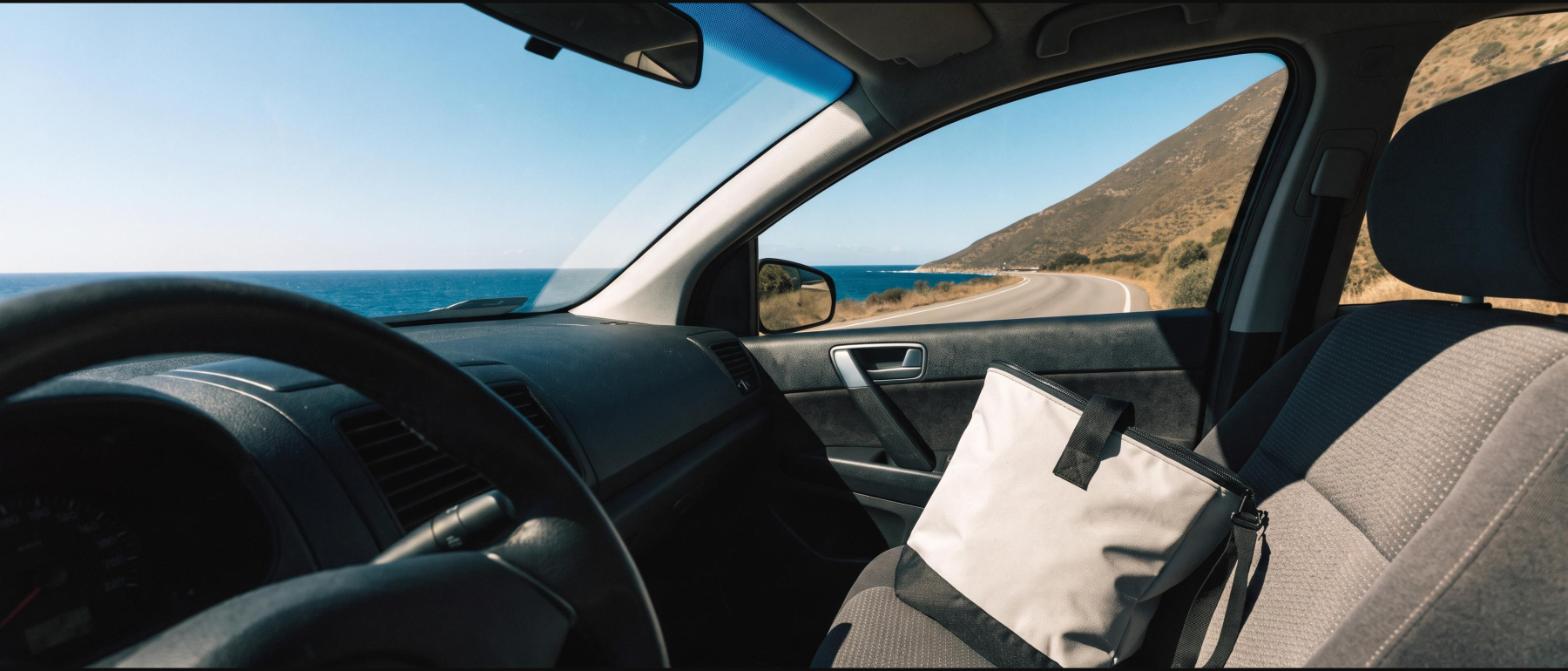 A soft insulated cooler bag on the passenger seat of a rental car on a coastal road